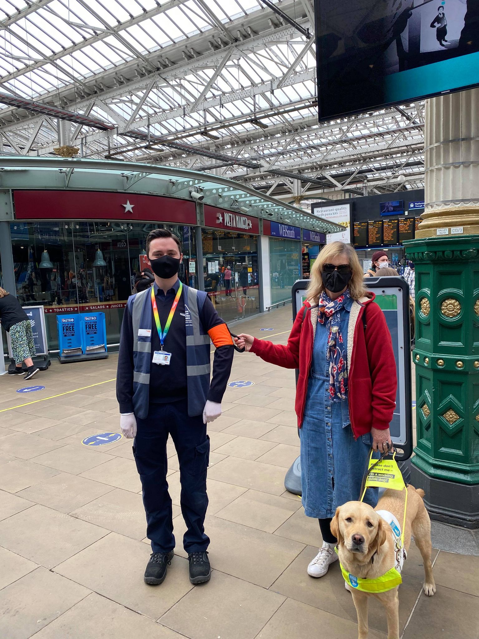 blind lady and her guide dog being guided by train staff using a ramble tag. they are in a train station